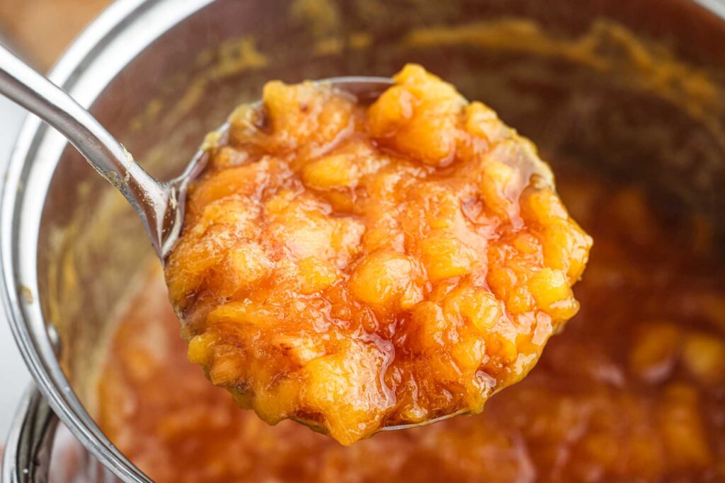 Large ladle lifting peach jam out of pot.